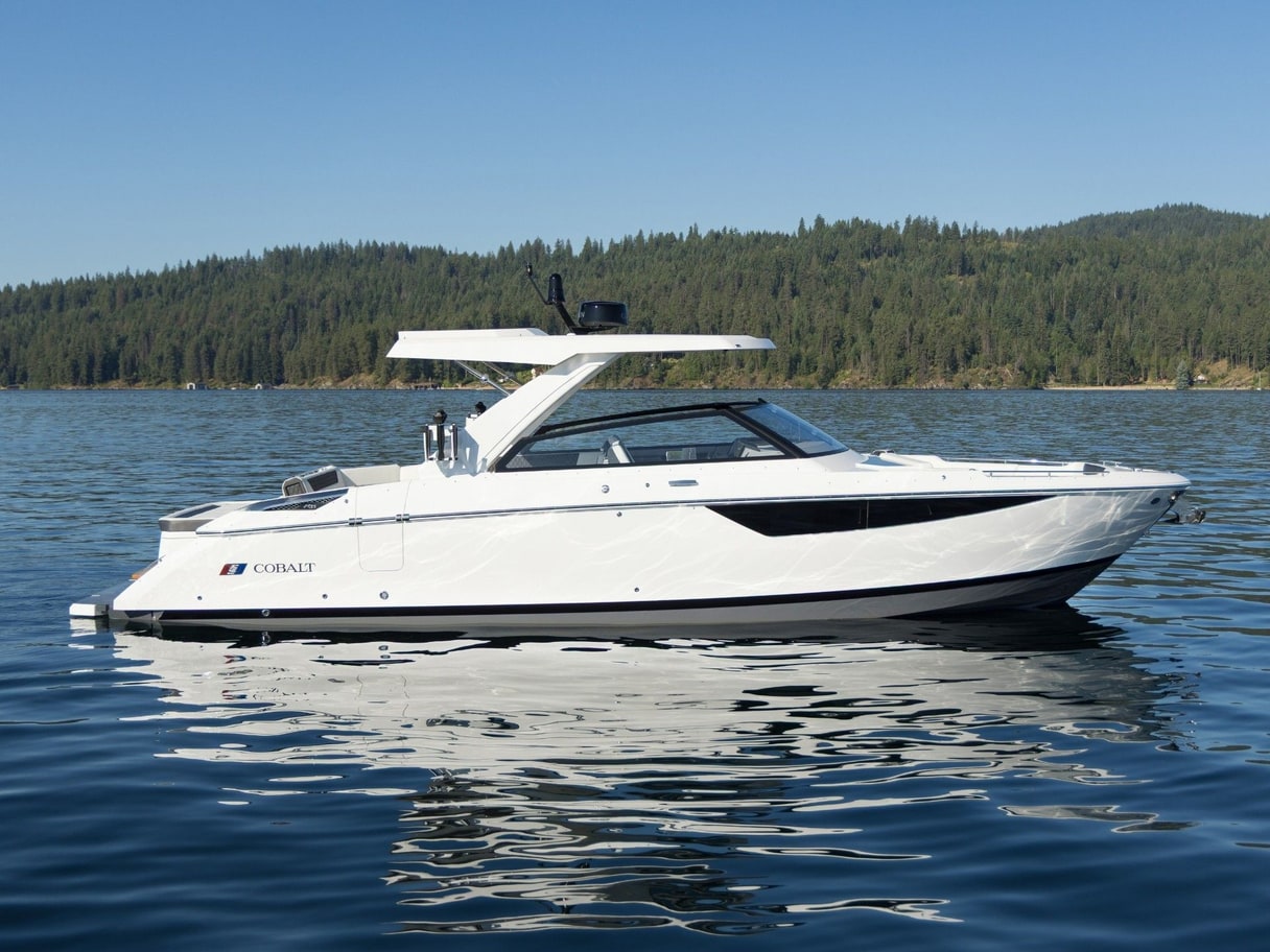 A white Cobalt cruiser on a calm lake in sunlight, with a tree-covered hillside and clear blue sky behind it.